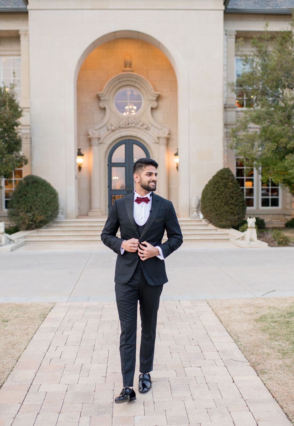 Groom in dark suit with burgundy bow tie stands before elegant stone arch building entrance on brick pathway.