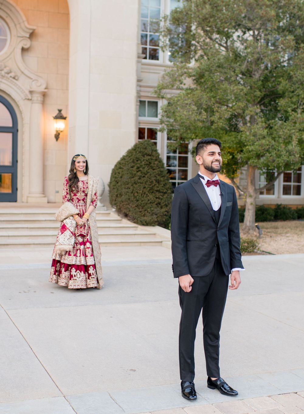 Groom in black tuxedo stands as bride in ornate red and gold Indian lehenga approaches during outdoor first look.