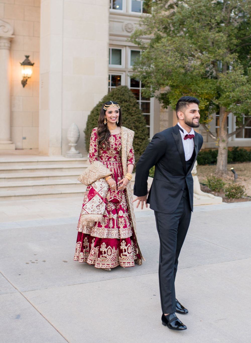 Indian bride in red lehenga and groom in black tuxedo walking together outside elegant venue.