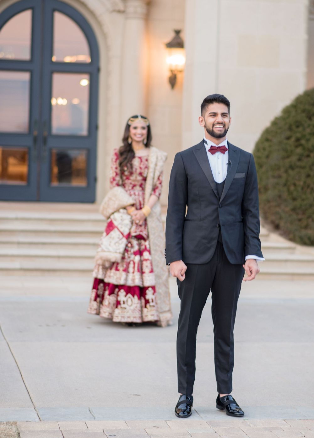 Groom in black suit smiling as Indian bride in red lehenga approaches grand venue entrance.