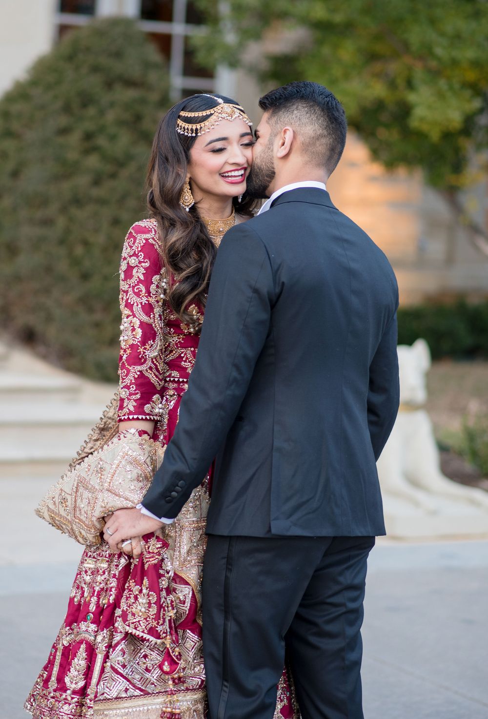 A couple in traditional South Asian wedding attire share a romantic moment, the bride smiling in a red and gold lehenga.