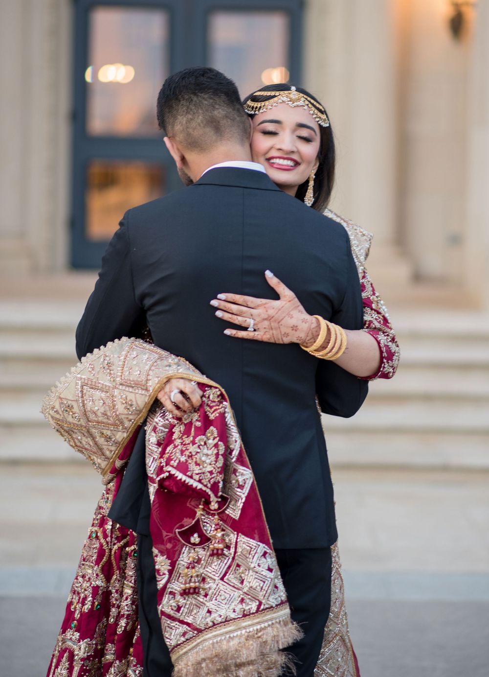 Bride in red and gold lehenga with maang tikka embraces groom in black suit at South Asian wedding ceremony.