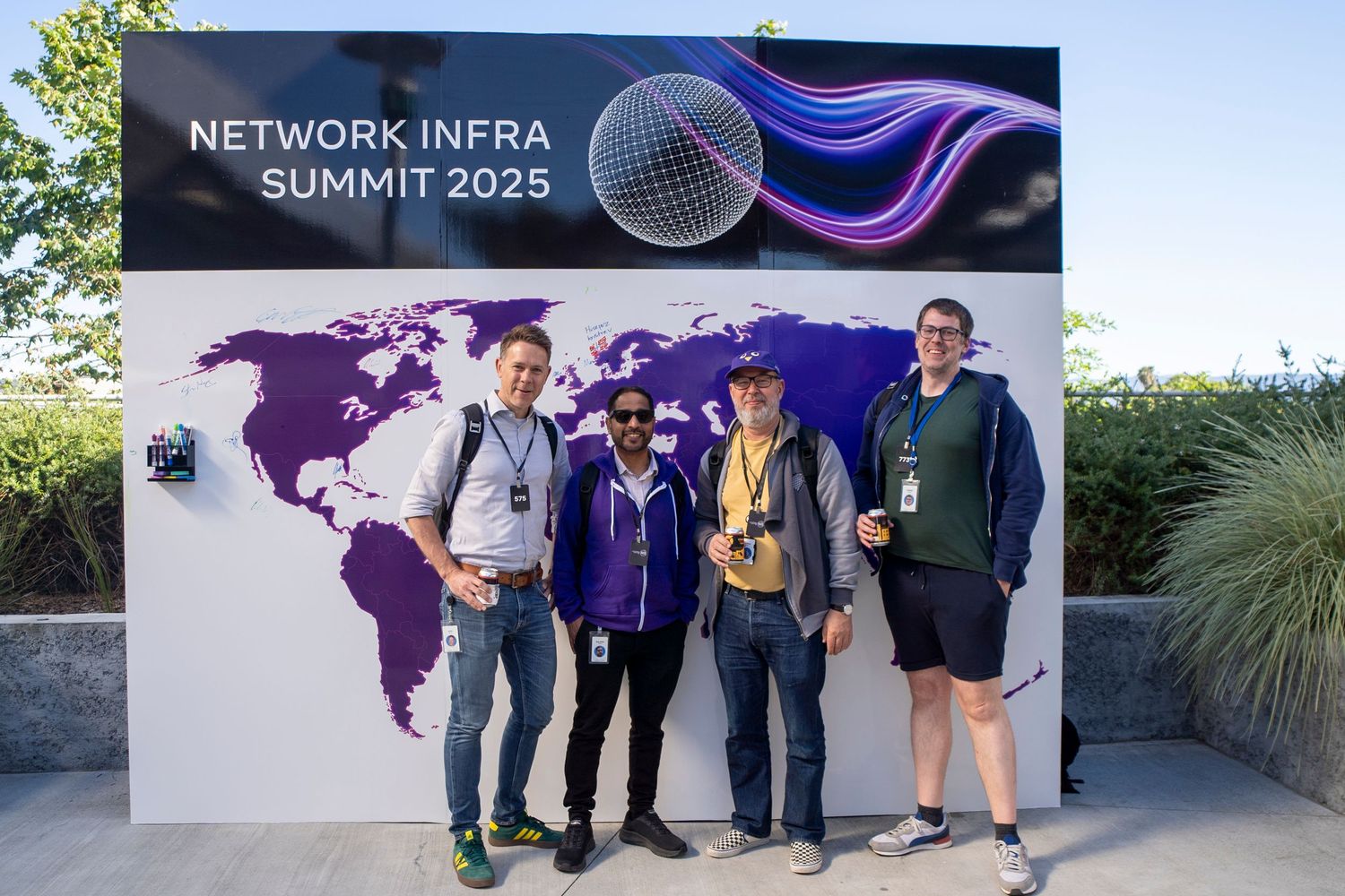Four attendees pose in front of the Network Infra Summit 2025 sign with a world map backdrop.