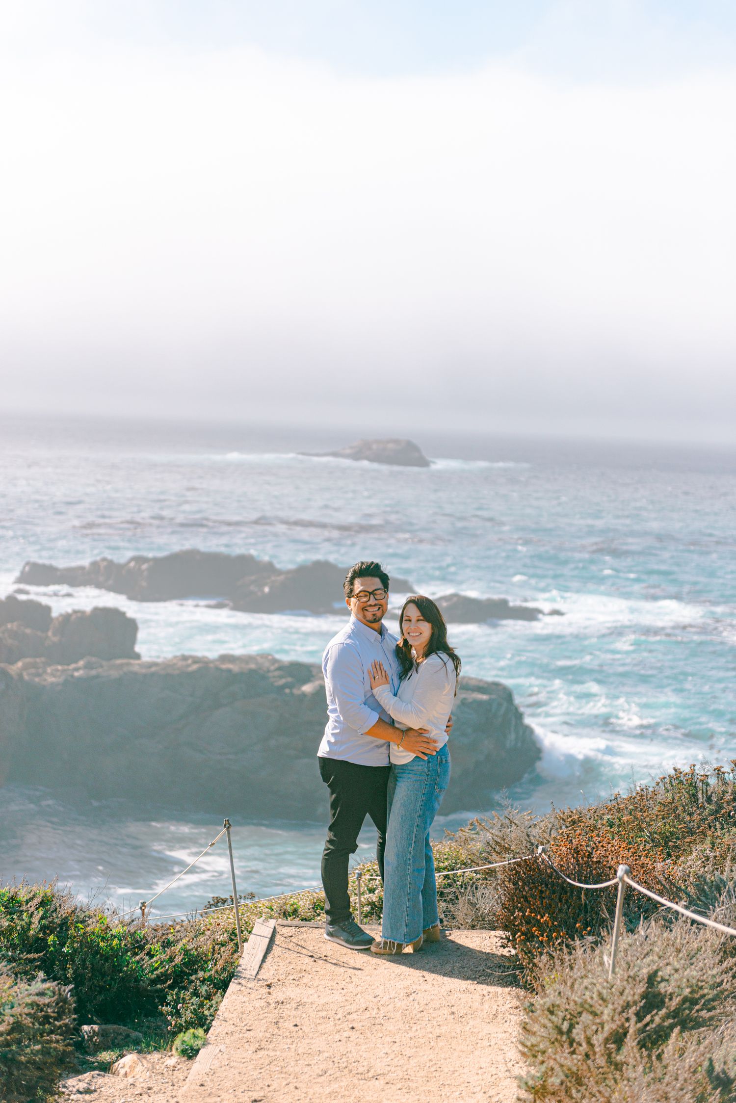 Couple embracing on a coastal cliff overlooking rocky shoreline and misty ocean waves at Big Sur, California.