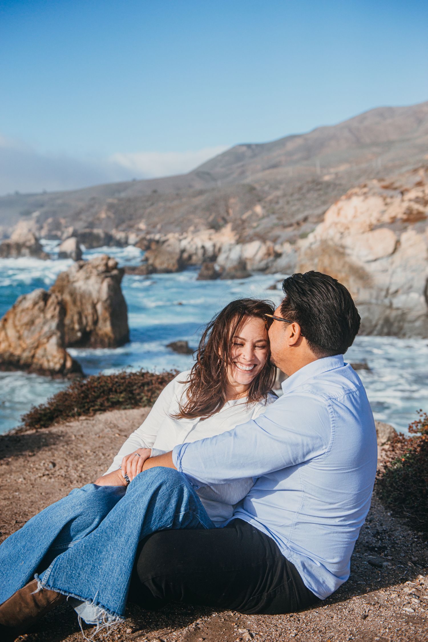 Couple sitting together on rocky coastal cliffs, smiling and embracing with ocean waves and mountains in the background.