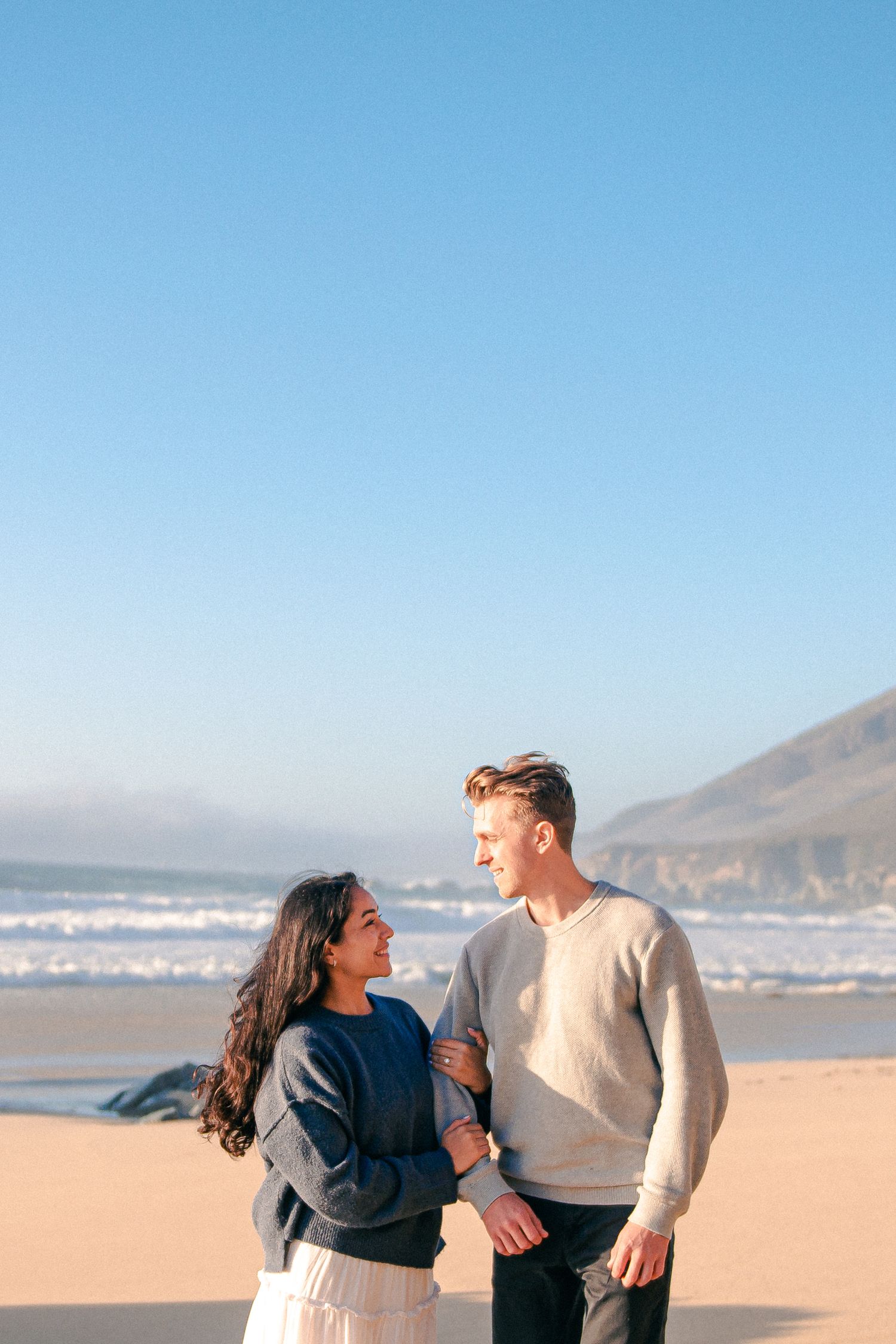 Couple standing together on a sandy beach, smiling at each other with ocean waves and cliffs in the background.