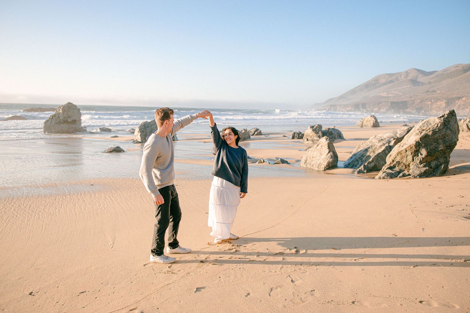 Couple dancing joyfully on a sandy beach with ocean waves and rocky coastline in the background.