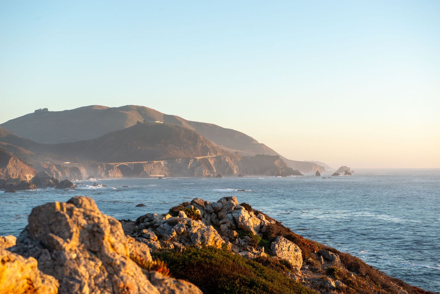 Rocky coastal cliffs overlook the Pacific Ocean at Big Sur, California, with a historic bridge and mountains at golden hour.