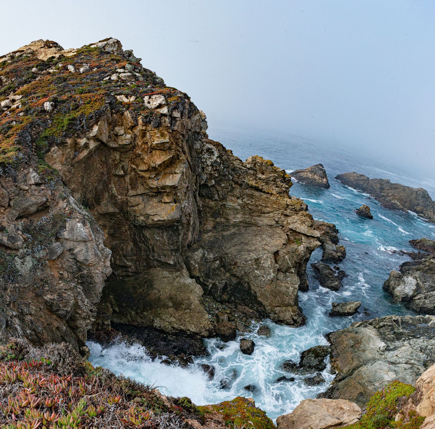 Rugged coastal cliffs with turquoise waves crashing against rocks in misty fog along a scenic shoreline.