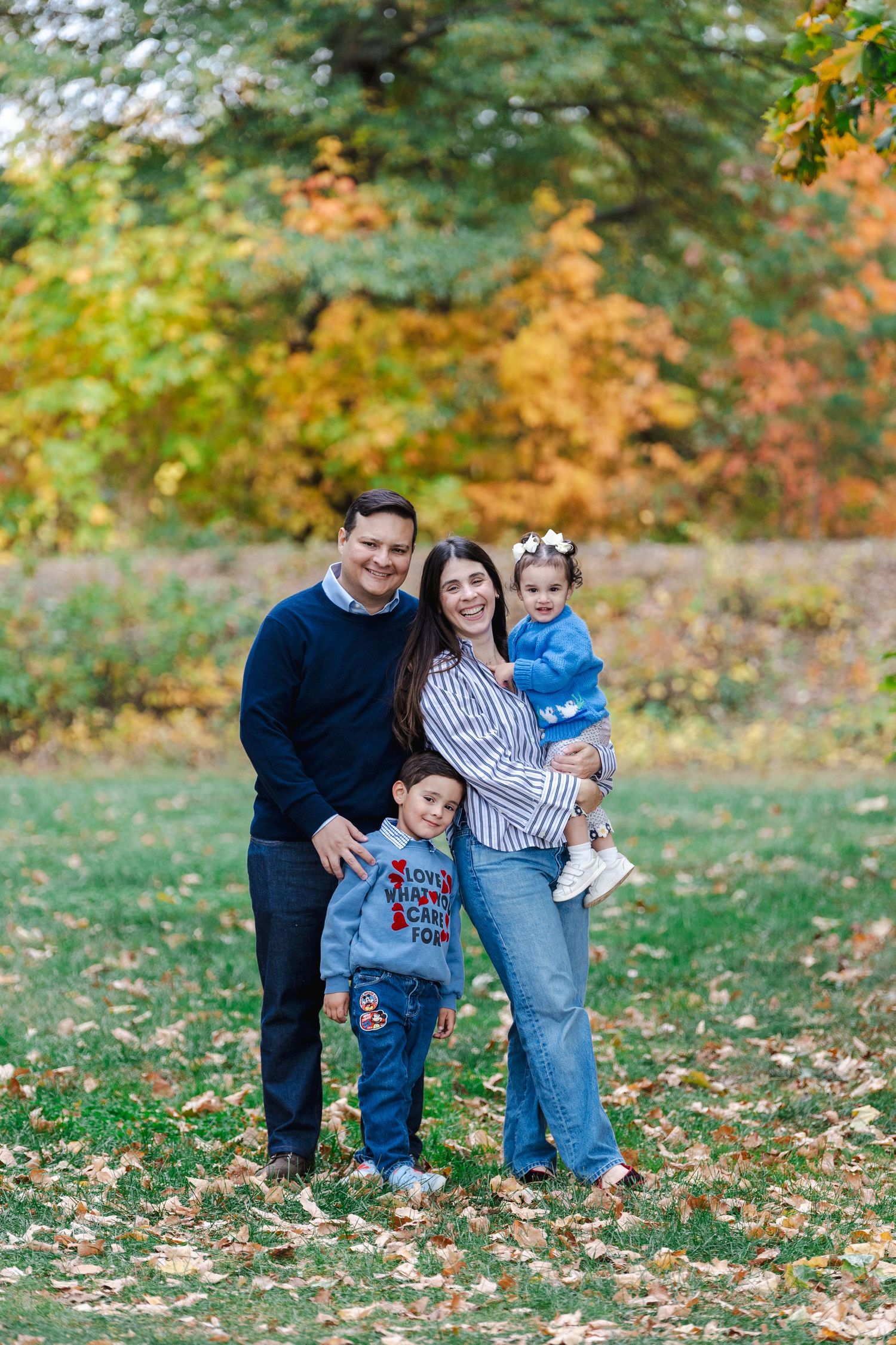 Family of four smiling outdoors in autumn park with colorful fall foliage, parents holding young children in casual attire.