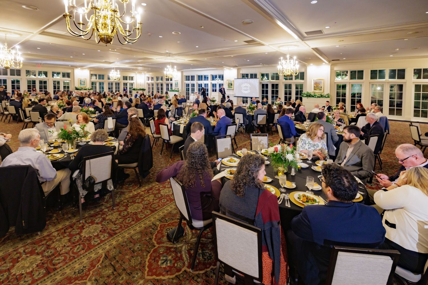 Elegant banquet hall filled with guests dining at round tables during a formal gala event with chandelier lighting.