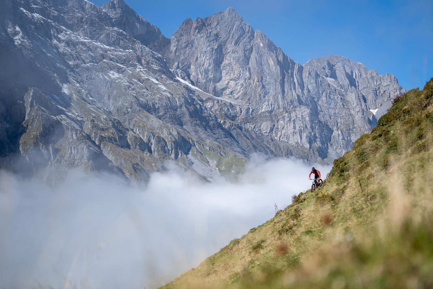 Mountain trail cuts across steep alpine slope with dramatic rocky peaks rising through clouds.