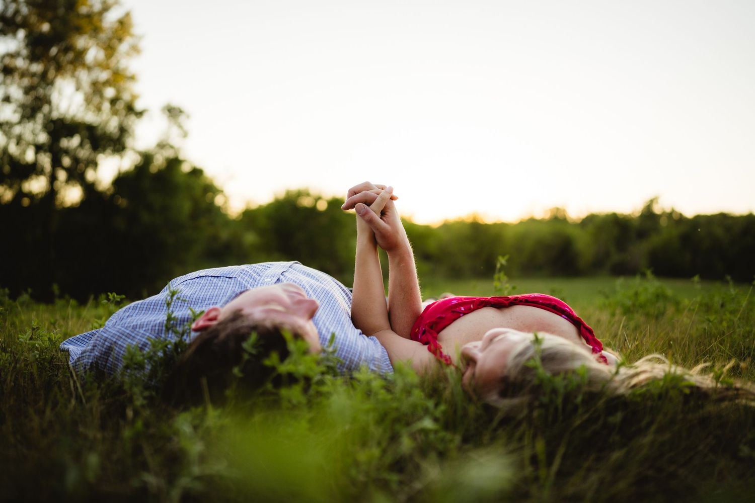 Two people lying on green grass holding hands at sunset in a romantic outdoor field setting.