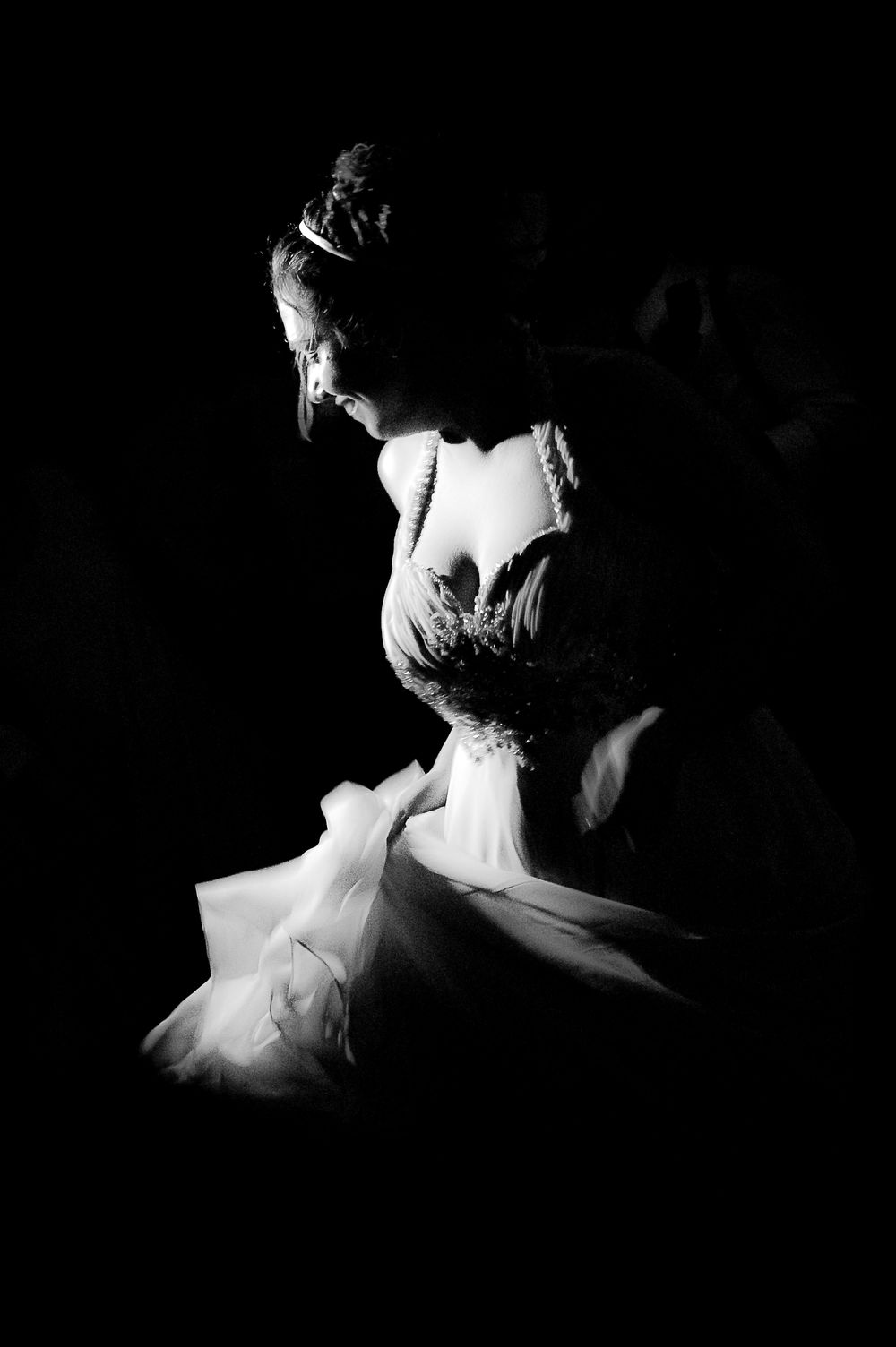 Elegant bride in a stunning wedding gown captured in dramatic black and white lighting, looking down gracefully.