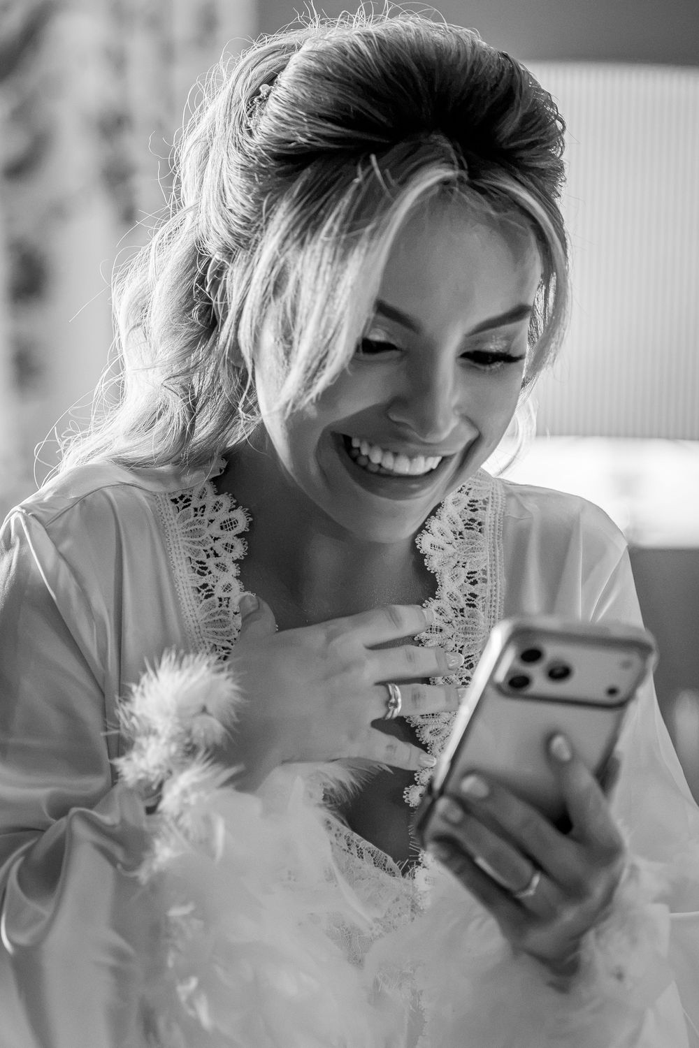 Black and white photo of a smiling bride in a robe holding a smartphone on her wedding day.
