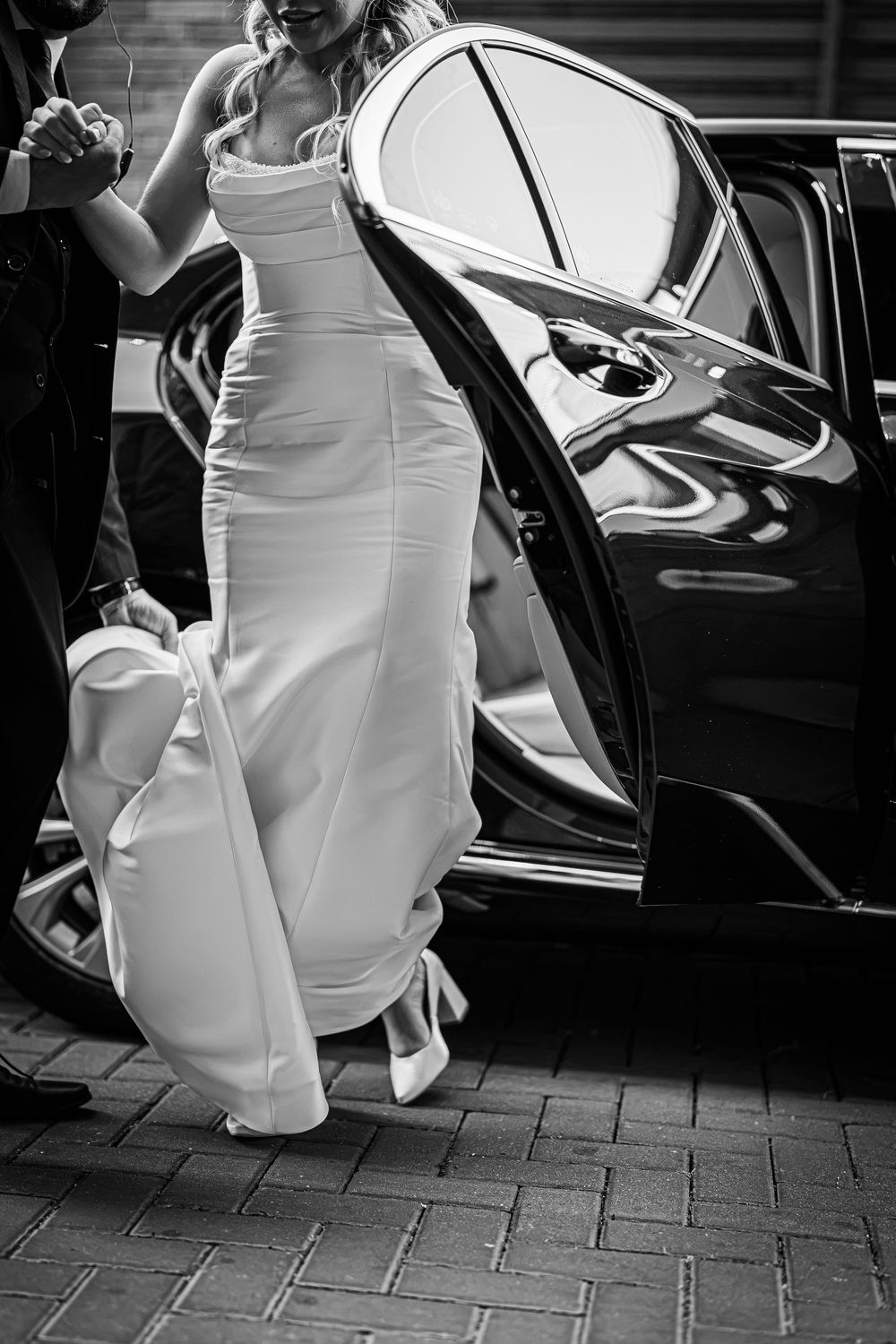 Black and white photo of a bride in a elegant white gown stepping out of a luxury black car.
