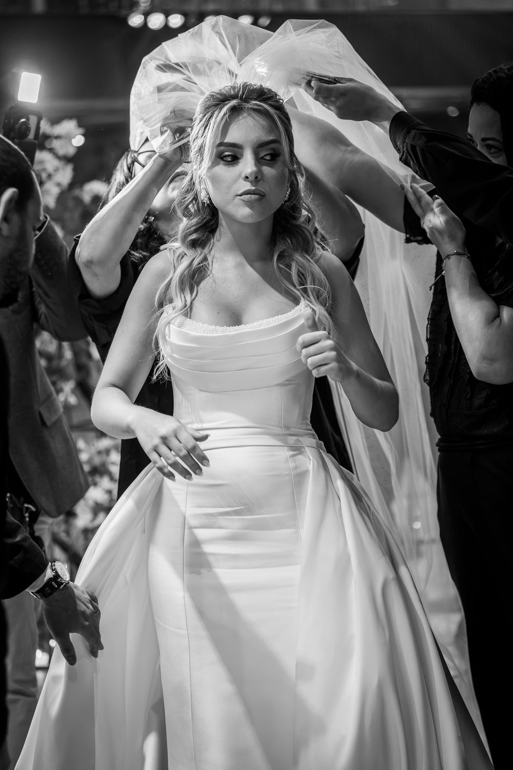 Black and white photo of a bride in a strapless white gown with veil being arranged by attendants.