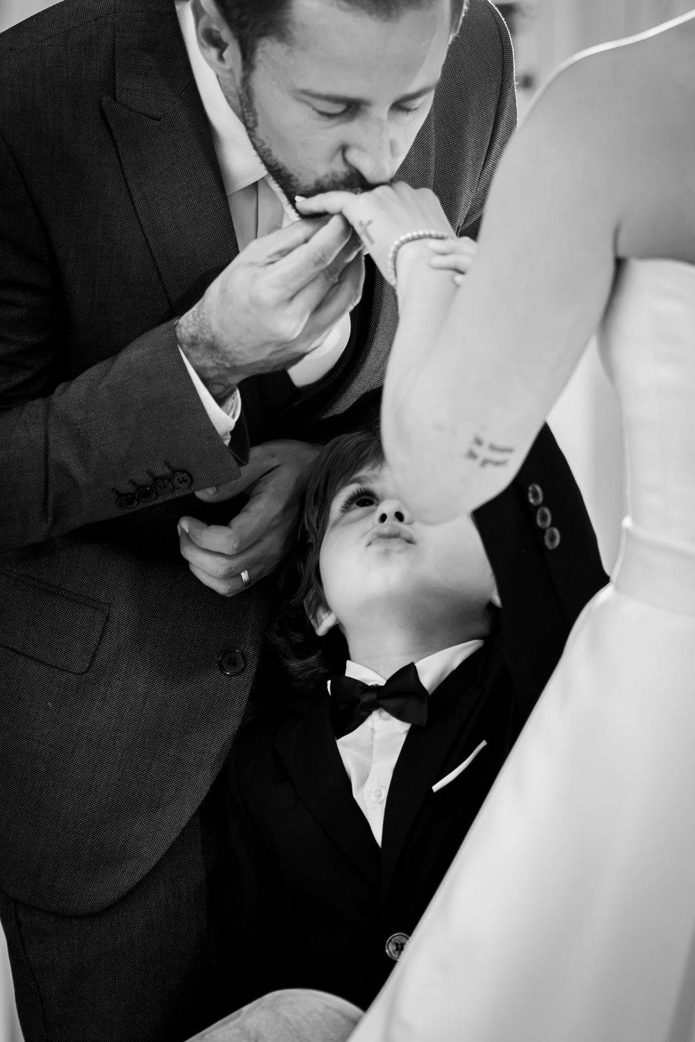 Groom in suit kisses bride's hand while young boy in tuxedo looks up during wedding ceremony, black and white photo.