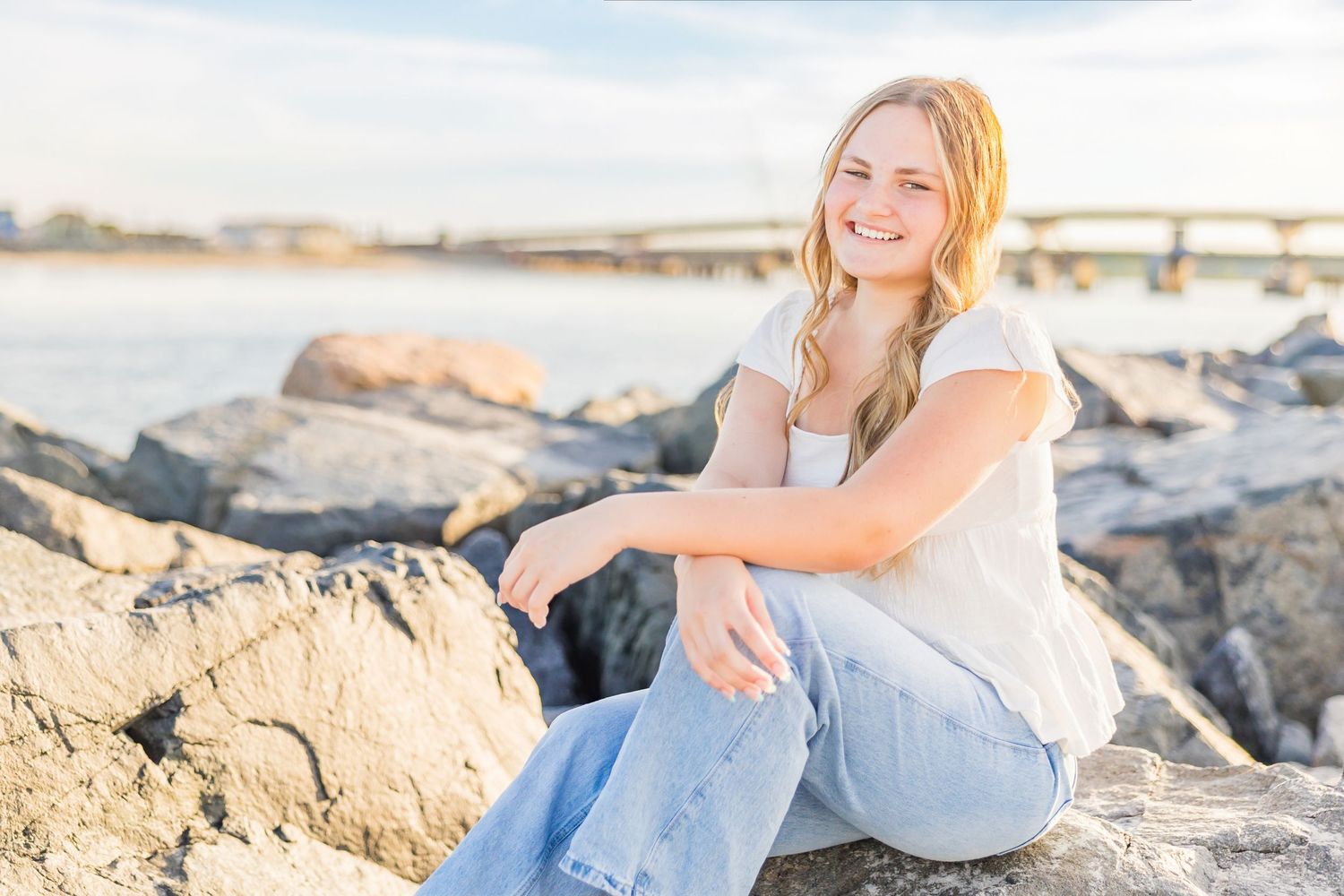 A smiling teenage girl sits on rocky shoreline in jeans and white top with a bridge and water in the background.