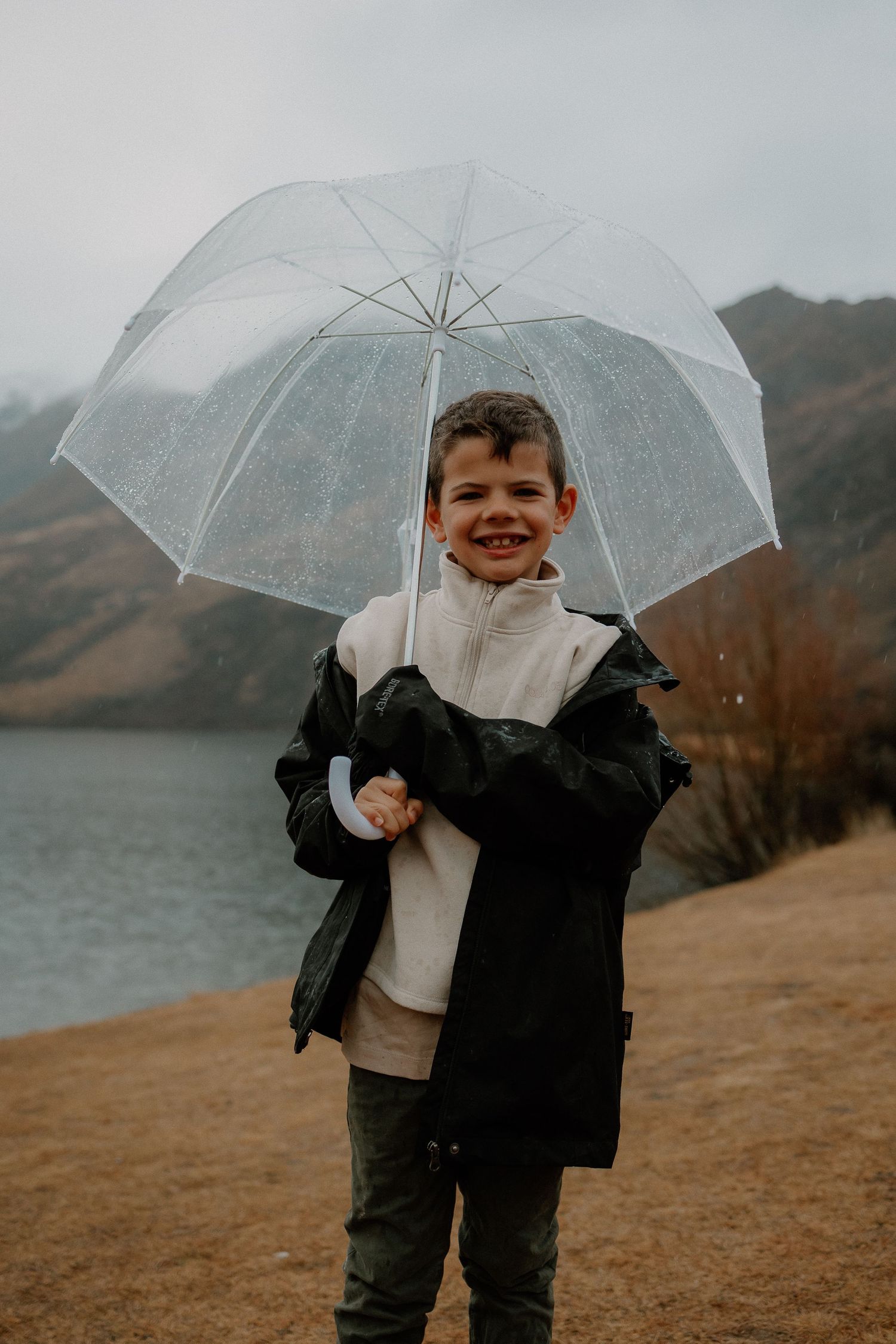 Smiling boy holding a clear umbrella in the rain near a lake, wearing a black jacket and cream sweater.