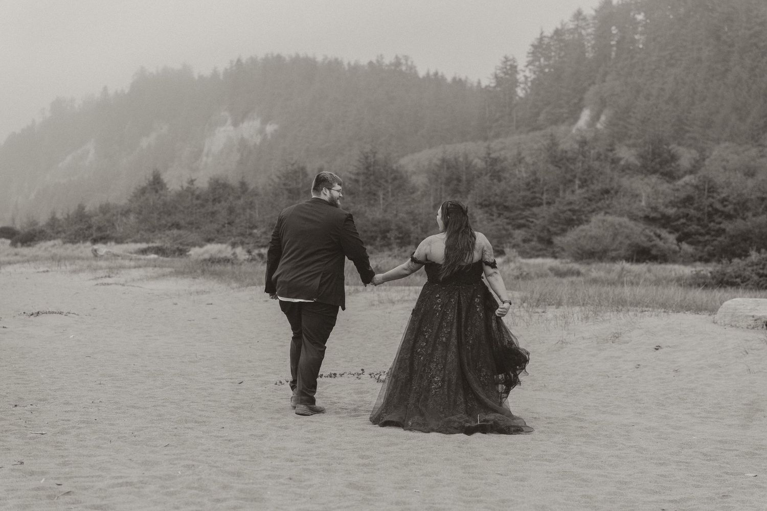Couple sharing a romantic kiss on a misty beach, woman in black flowing gown, surrounded by lush green hills.