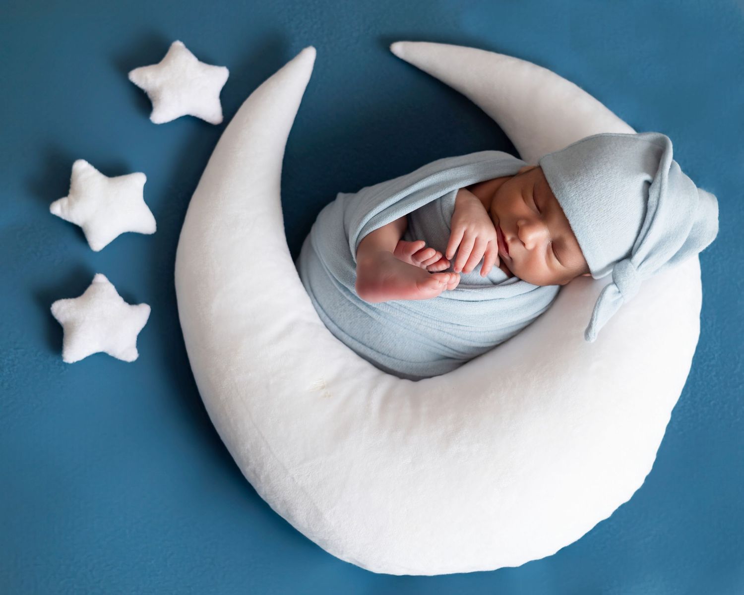Newborn baby sleeping peacefully on a crescent moon pillow with star props on a blue background.