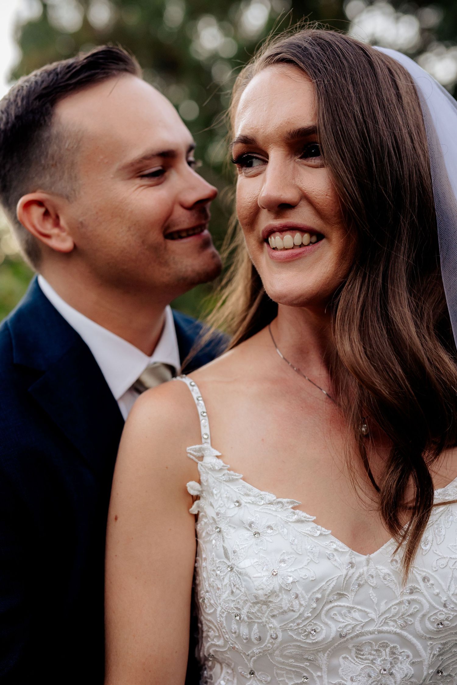 A smiling bride in a white lace gown and veil stands close with a groom in a navy suit at their wedding.