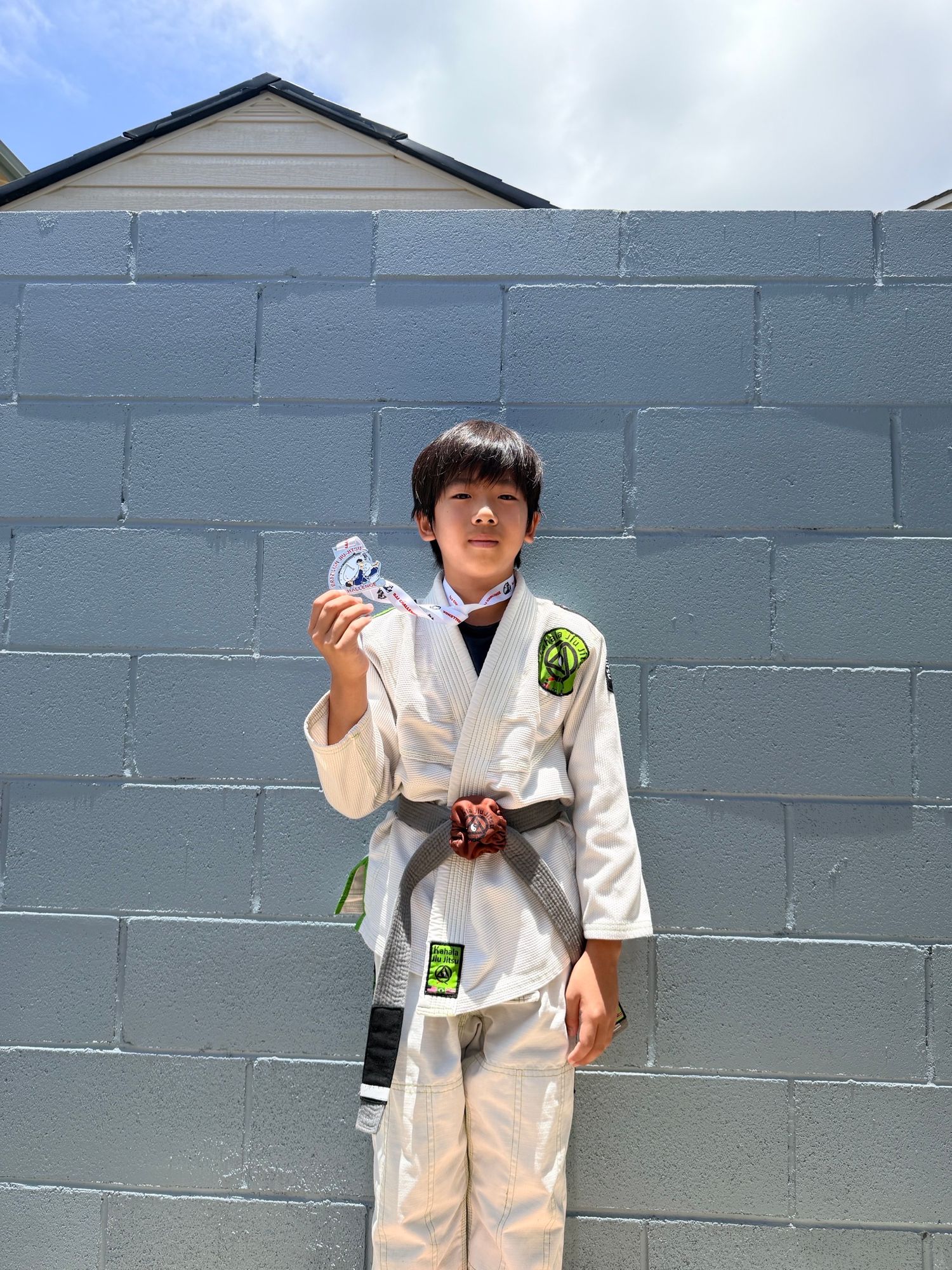 Young boy in white BJJ gi with gray belt holds a medal outdoors, standing proudly against a gray concrete block wall.
