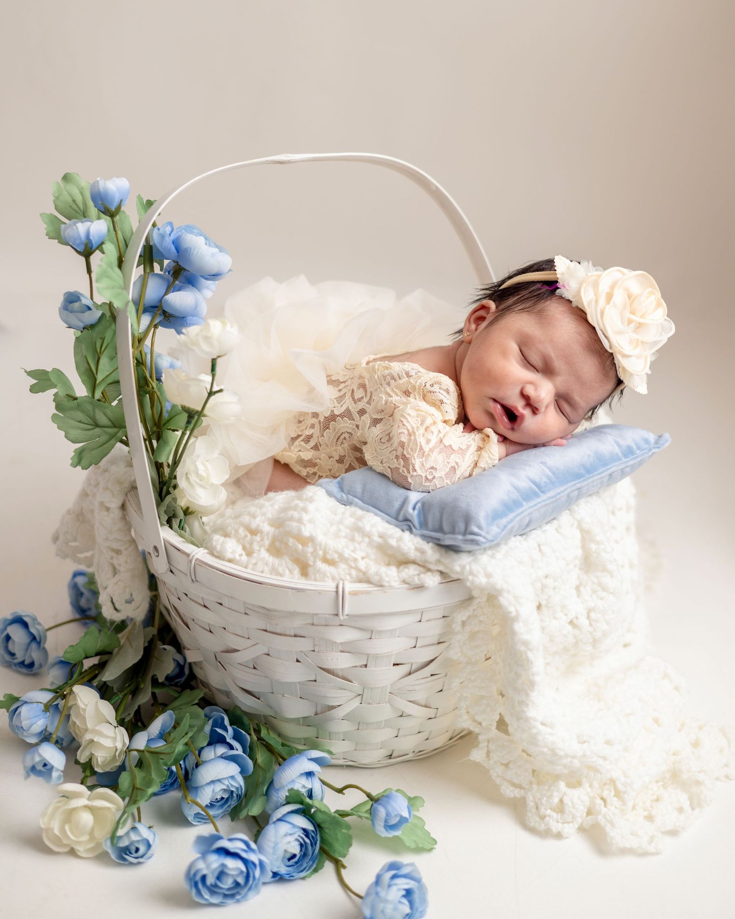 Sleeping newborn baby in lace outfit resting in white wicker basket with blue roses and cream blanket.