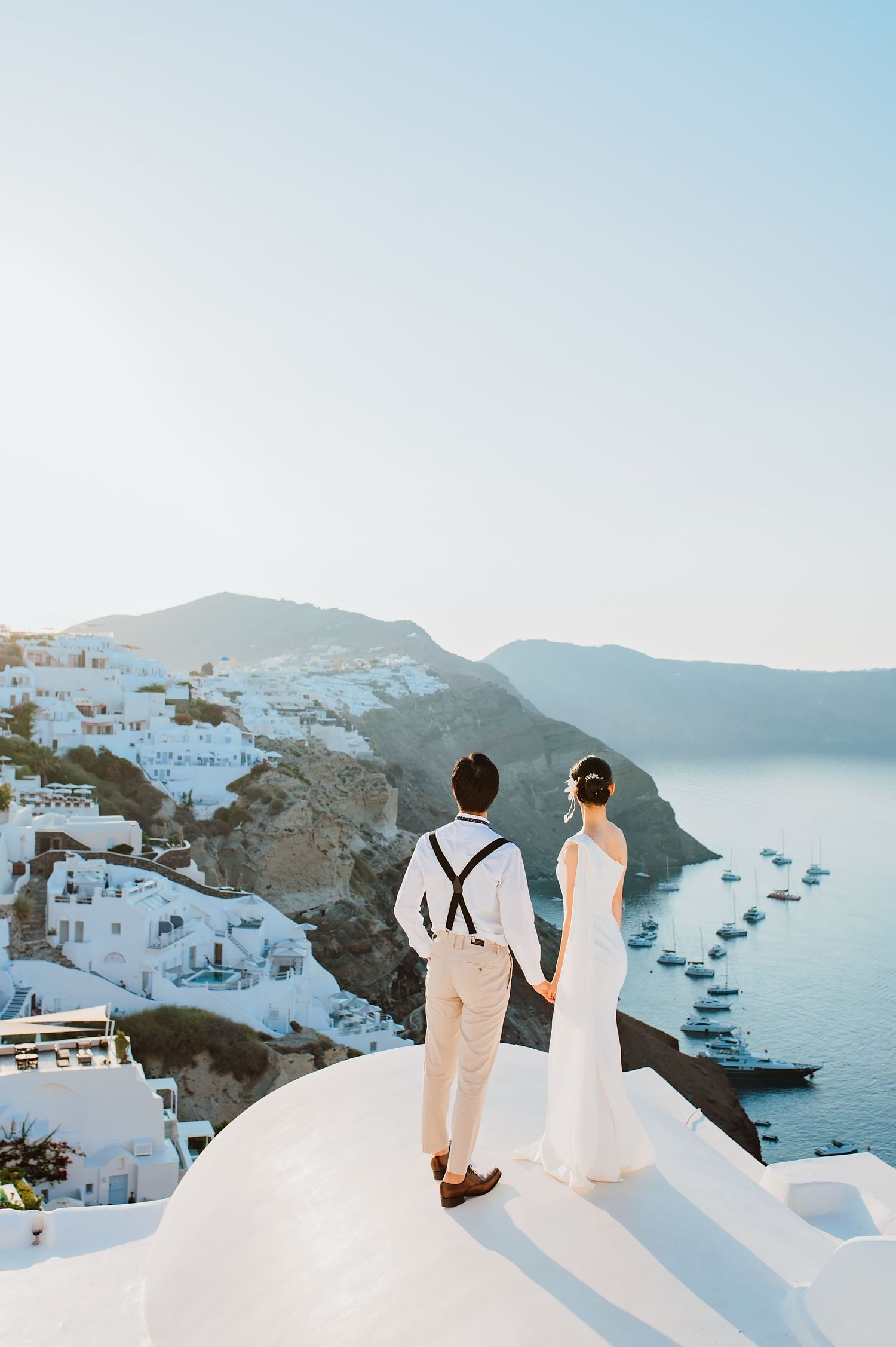 Couple in wedding attire holding hands on a rooftop overlooking Santorini caldera with white buildings and blue sea.