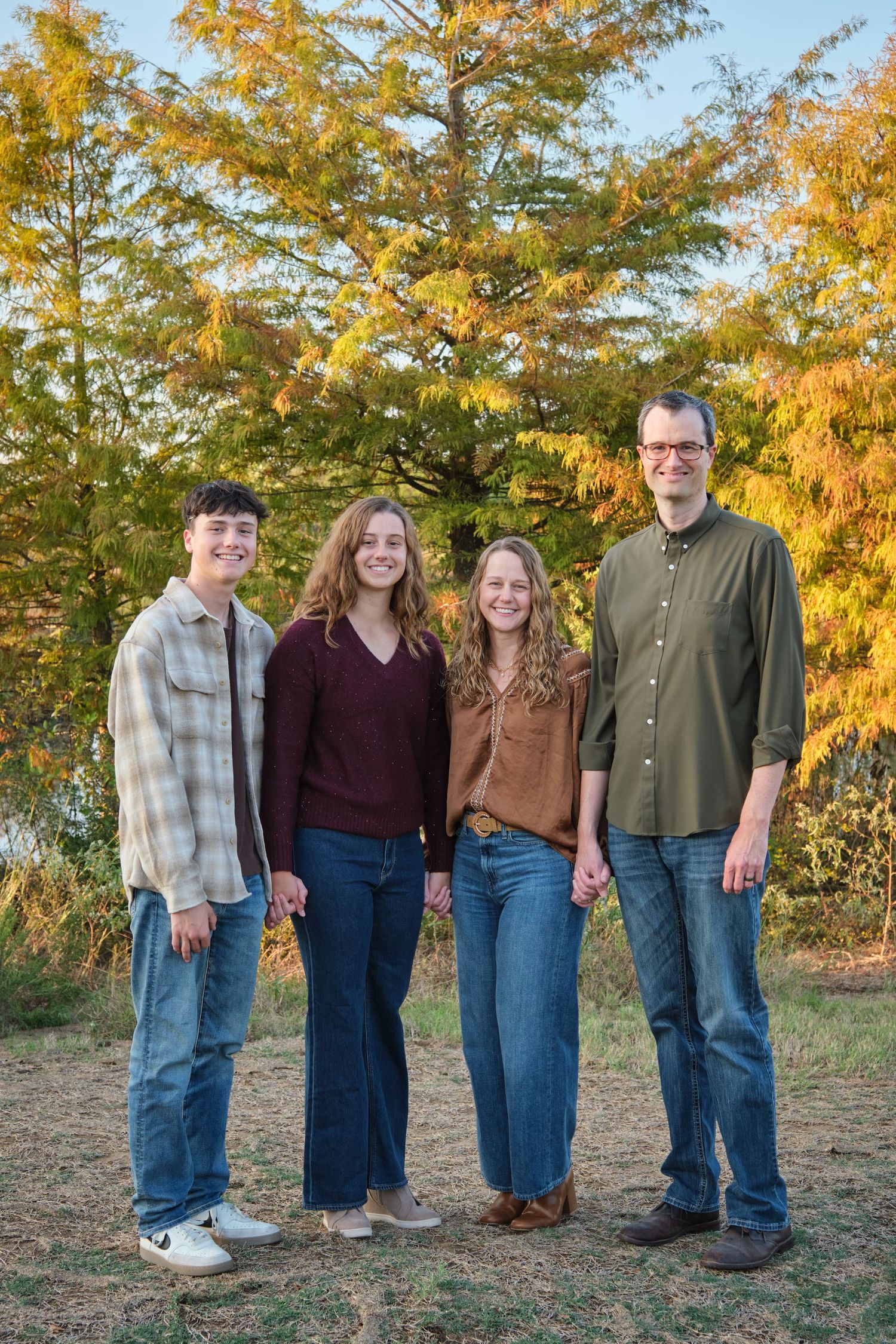 Family of four smiling outdoors in autumn, standing before colorful fall foliage trees on a sunny day.