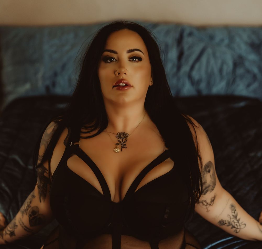 Woman with tattoos wearing black lingerie and rose necklace poses confidently on a dark couch.