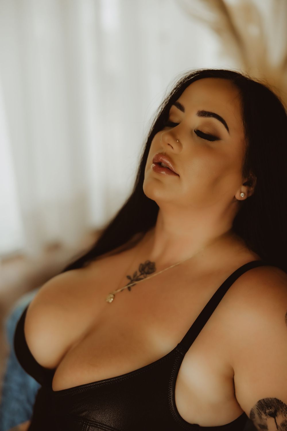 Woman with long dark hair in black leather top, eyes closed, wearing gold necklace and rose tattoo on chest.