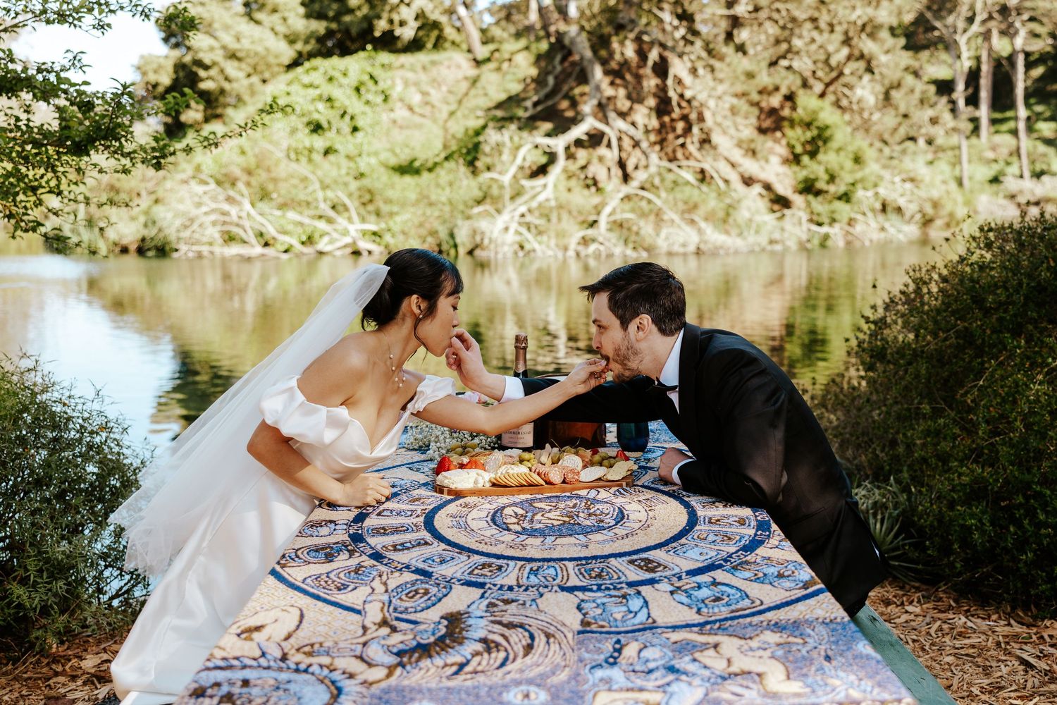 couple feeding each other charcuterie snacks in golden gate park with blue heron lake behind them during SF elopement day