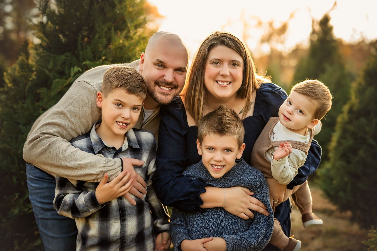 Happy family of five smiling outdoors near evergreen trees during golden hour in a warm, candid portrait.