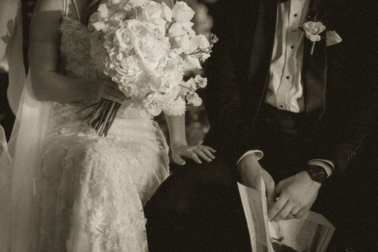 Black and white wedding photo of bride holding white bouquet beside groom in tuxedo during ceremony.