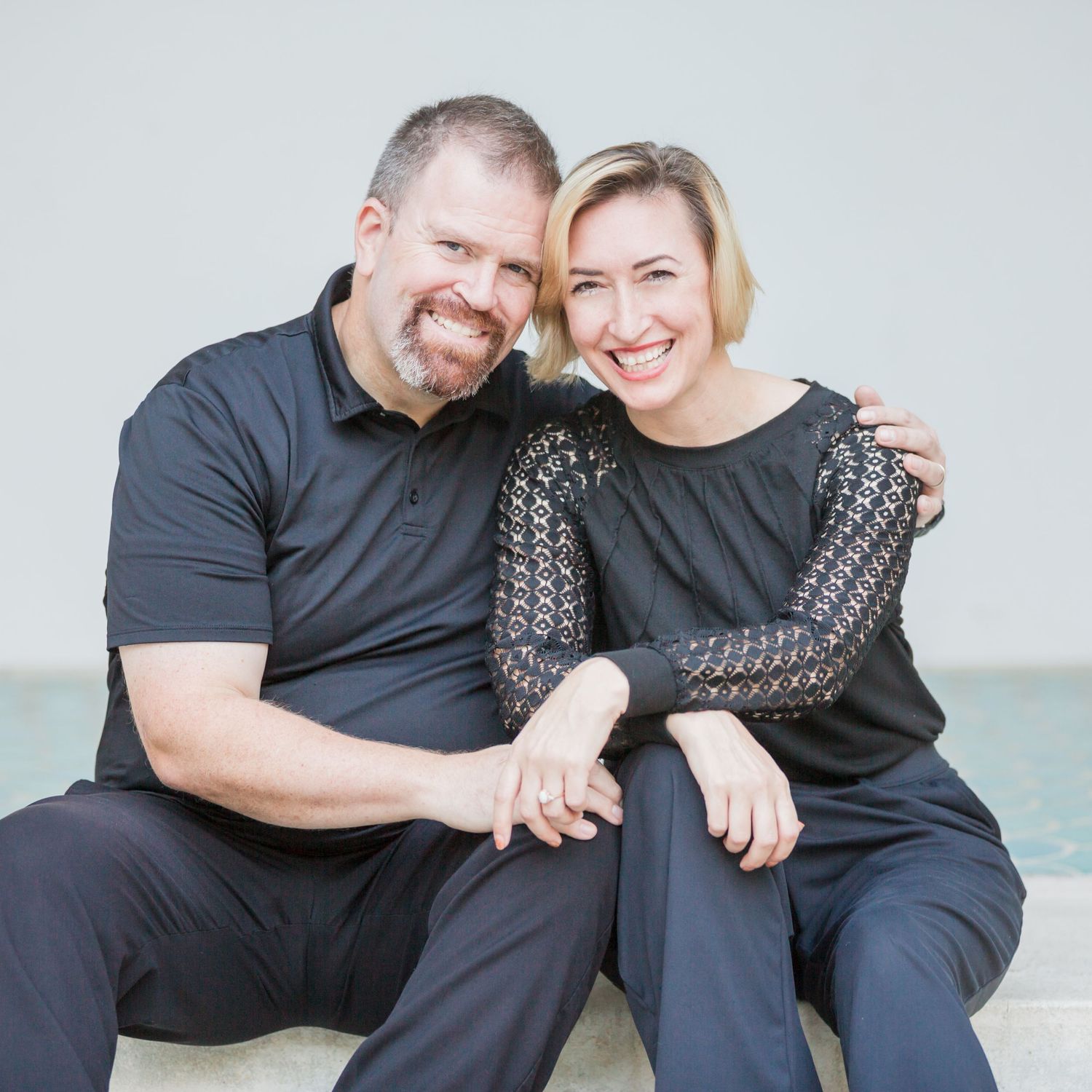 A smiling couple in matching black outfits sitting closely together, the man with his arm around the woman, against a light background.