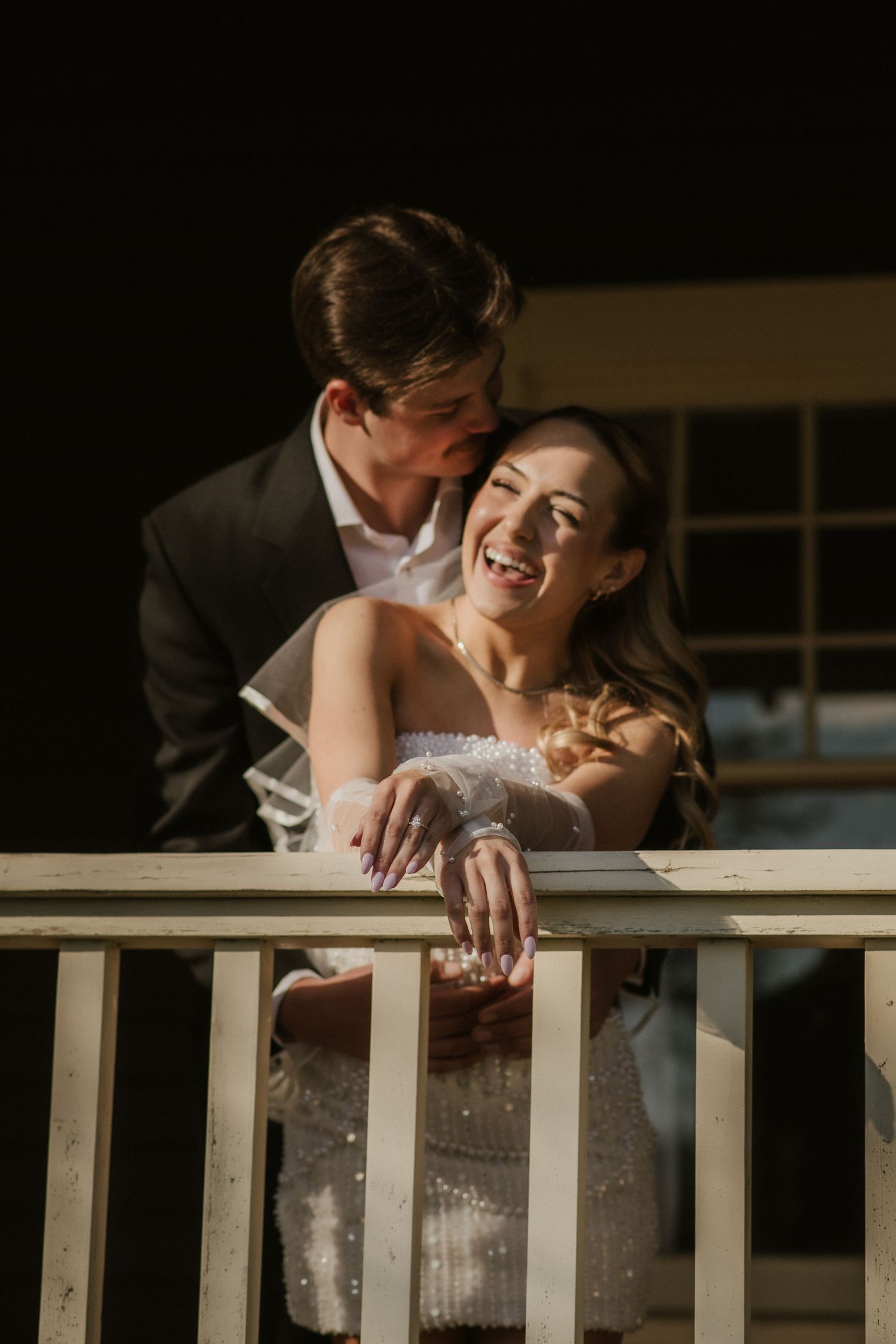 A joyful bride in a strapless gown laughs on a balcony as her groom in a dark suit kisses her cheek.