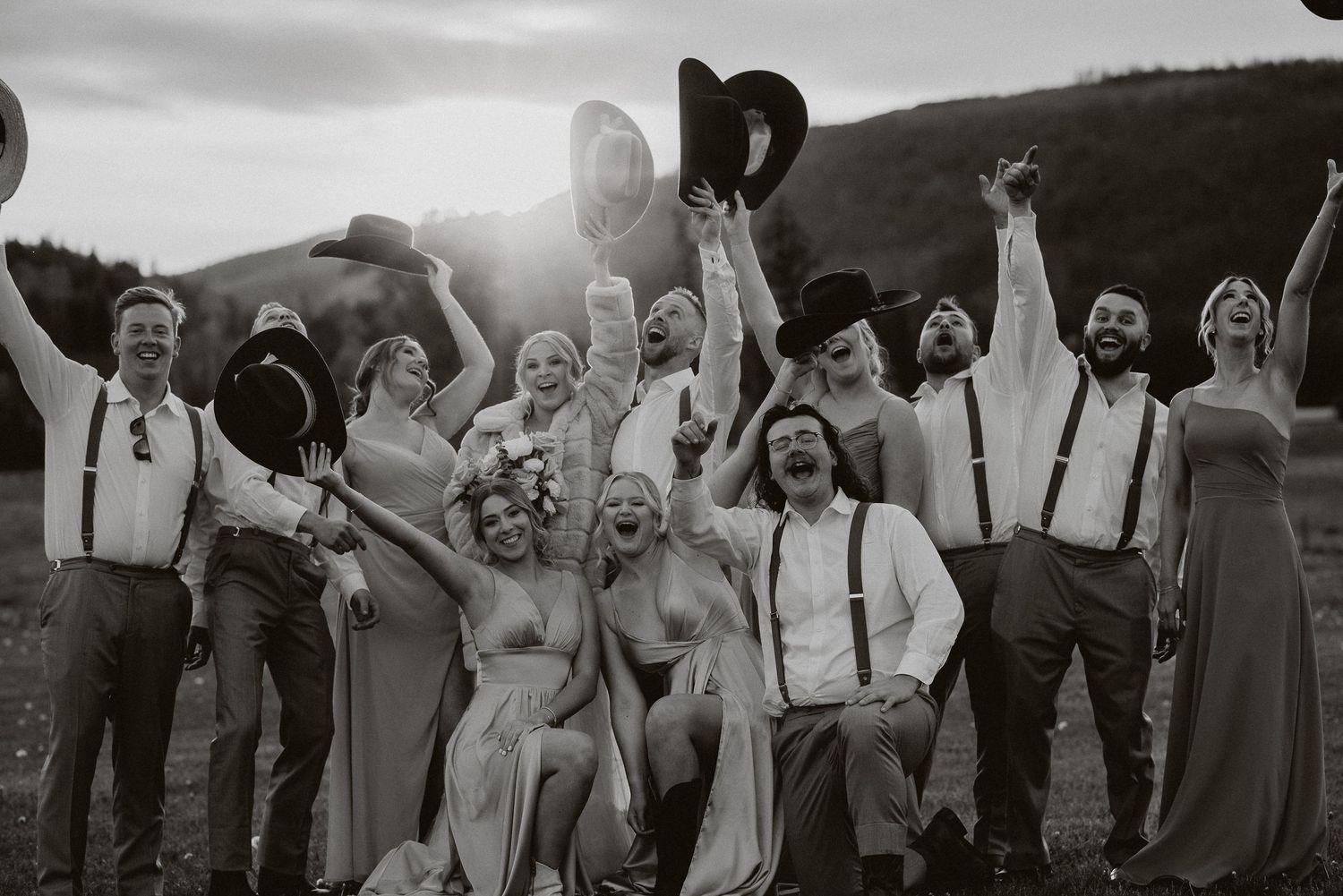Black and white photo of a joyful wedding party celebrating outdoors, raising hats and cheering in a scenic mountain setting.