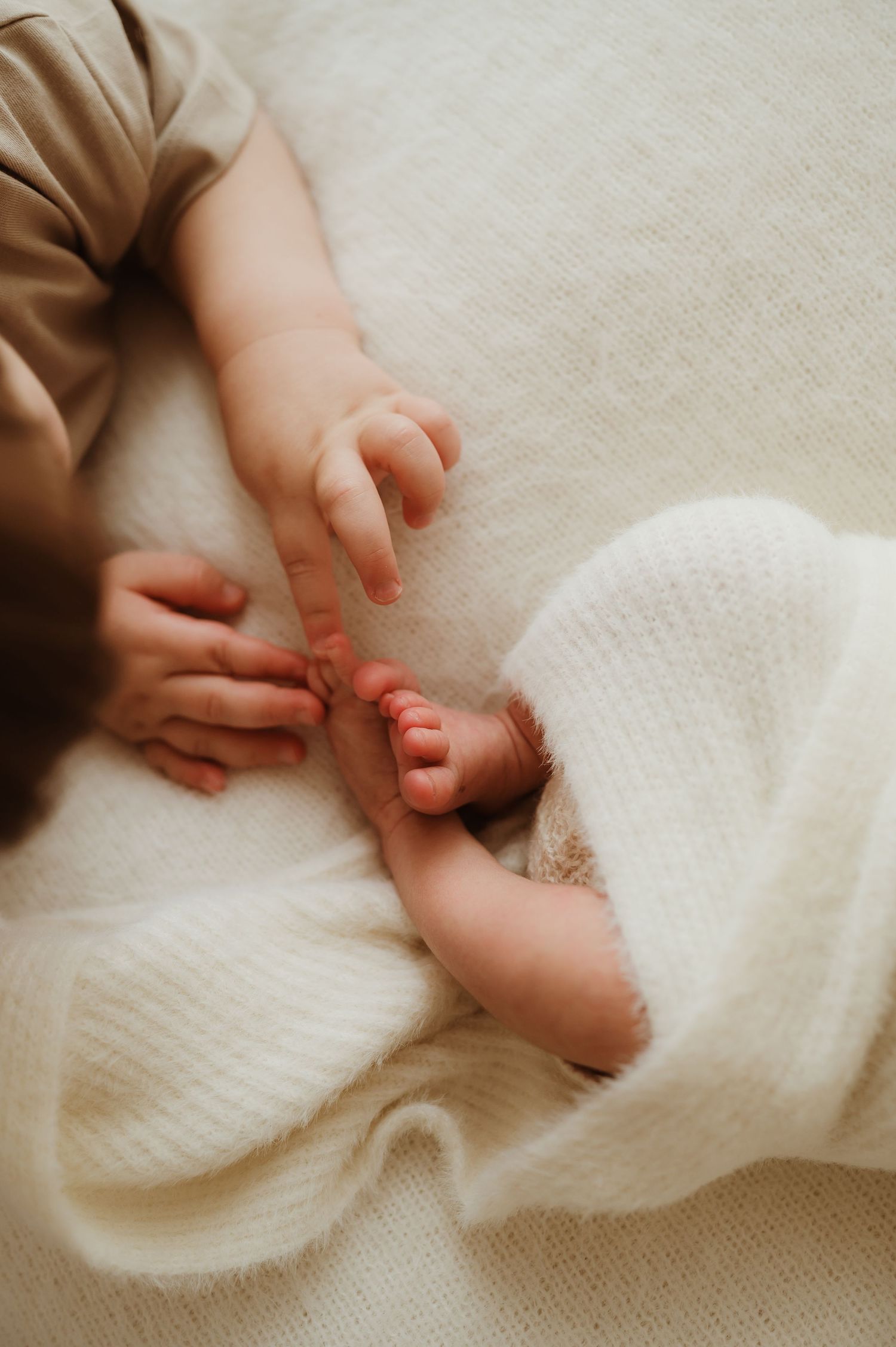 A toddler gently holds the tiny feet of a newborn baby wrapped in a soft white knit blanket.