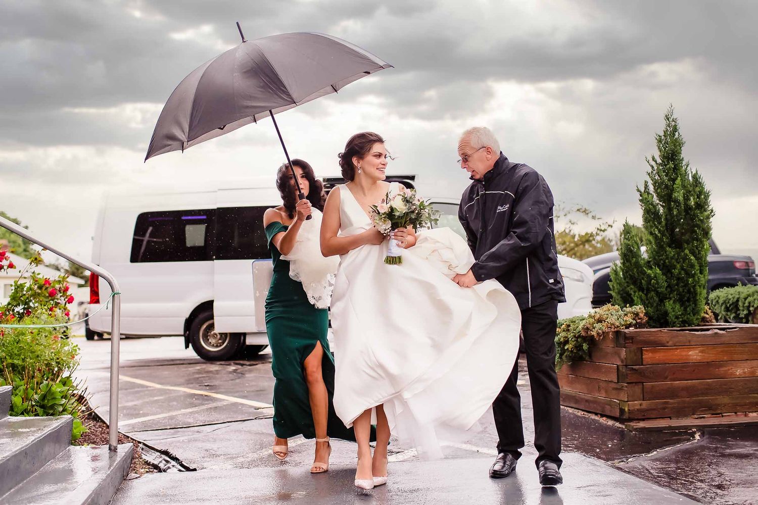Bride laughing in the rain while her father and bridesmaid rush to hold her dress and an umbrella outside the venue.