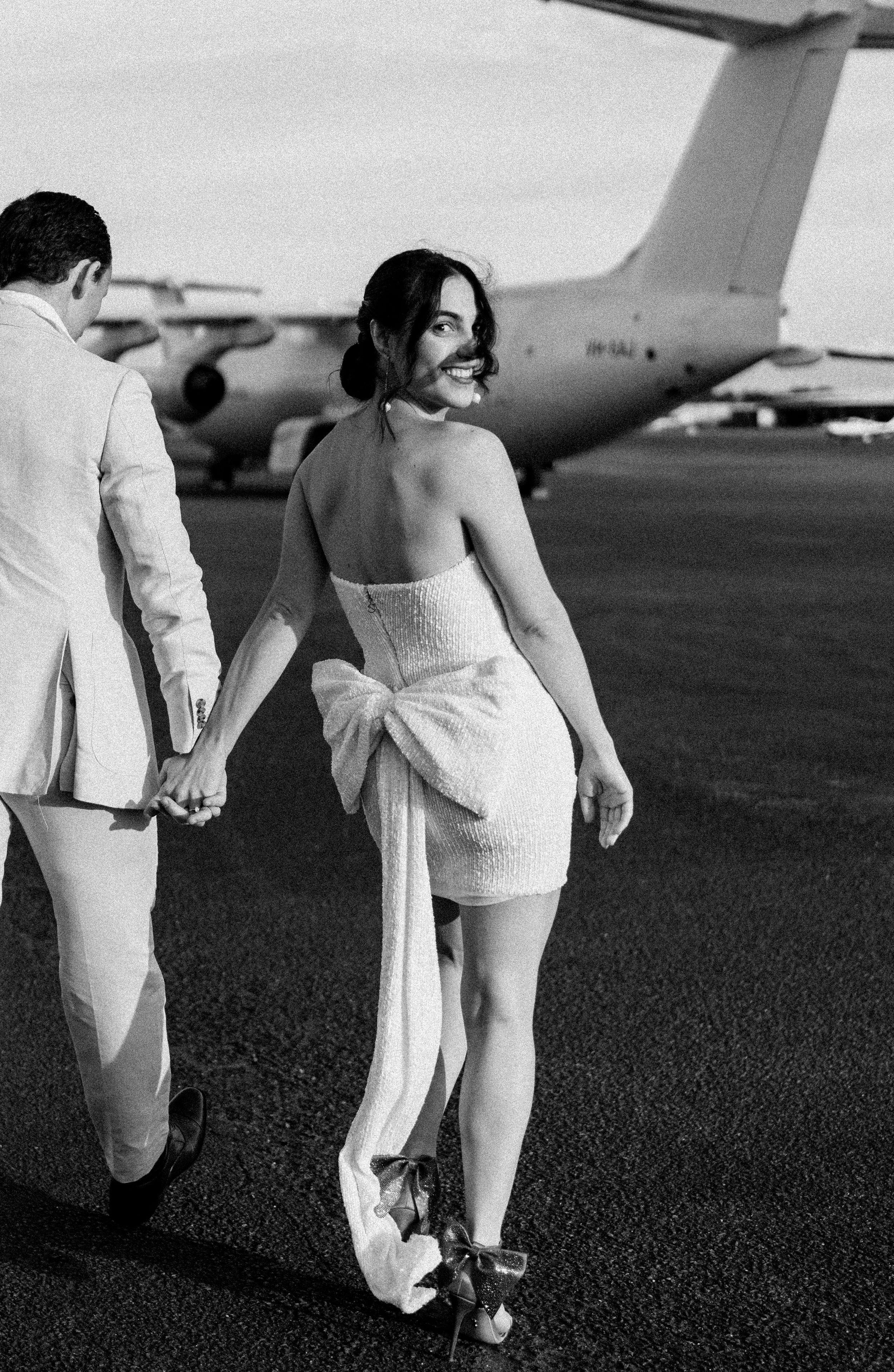 A couple in wedding attire holds hands on an airport tarmac, bride in backless mini dress with bow, groom in white suit.