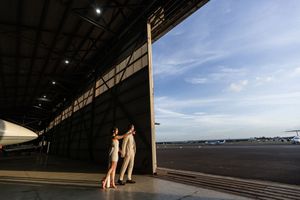 Two elegantly dressed women stand in a large aircraft hangar doorway overlooking a sunny airport runway.