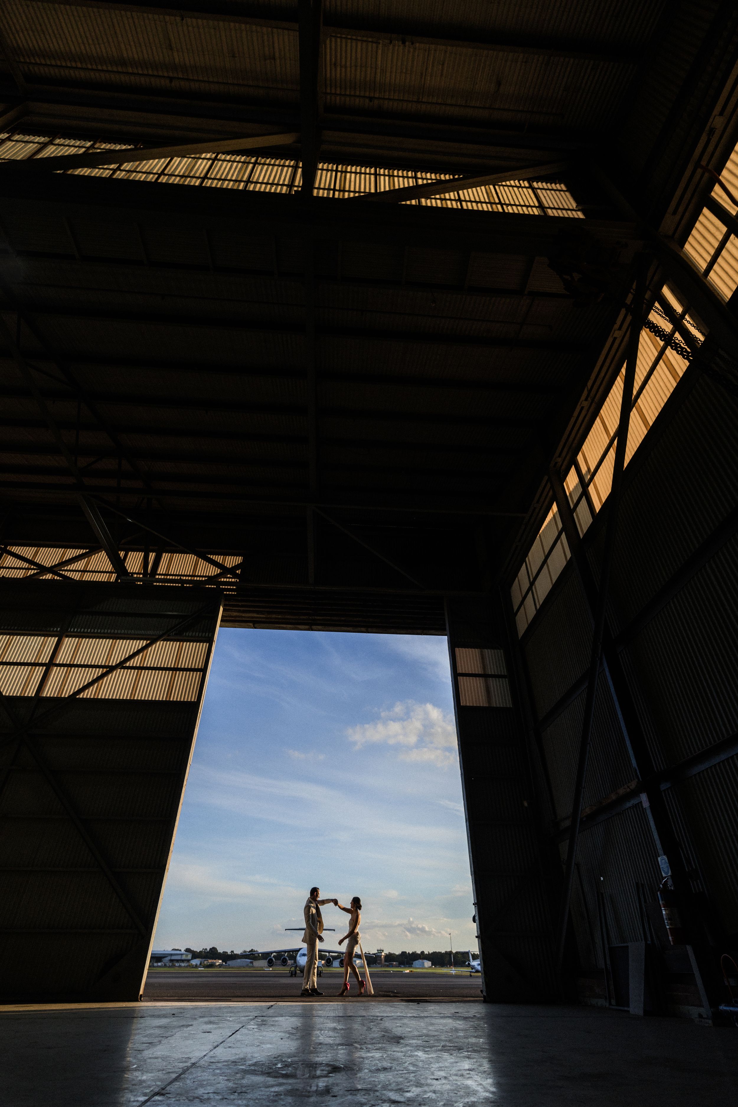 Silhouette of two people holding hands in a large aircraft hangar doorway with blue sky backdrop.
