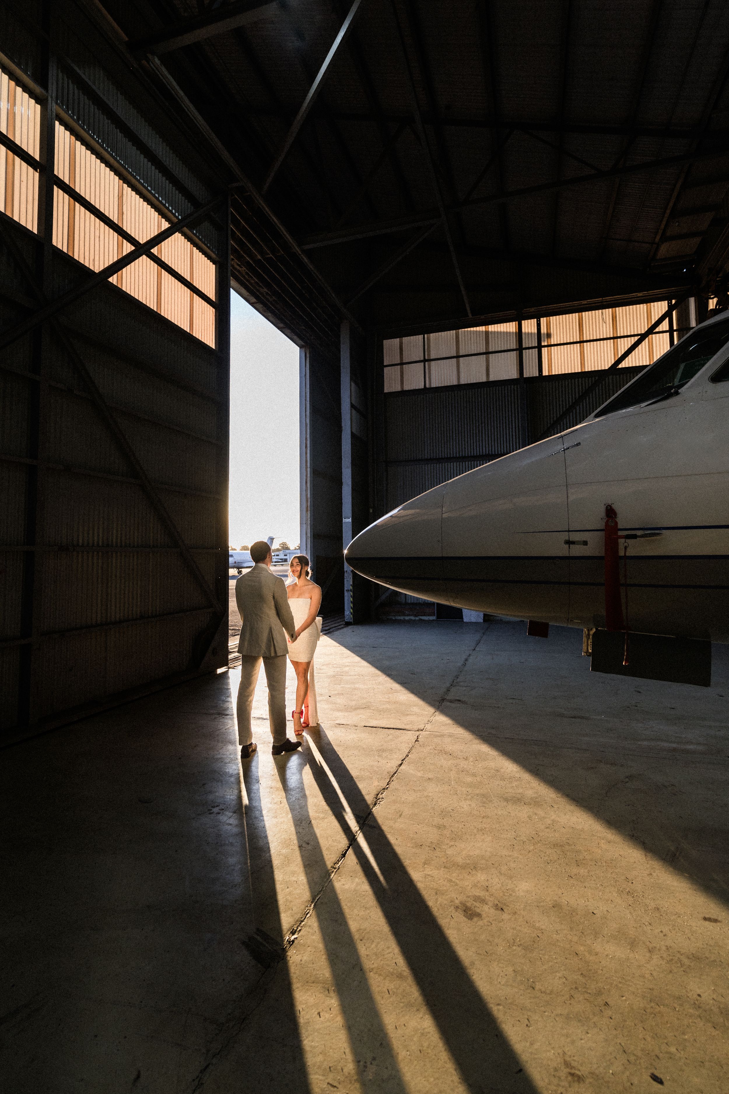 Couple holding hands in a sunlit airplane hangar with a private jet, casting long shadows on the concrete floor.