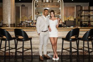 Elegant couple posing at a stylish bar, man in white suit, woman in short white dress, perfect for wedding or event venue.