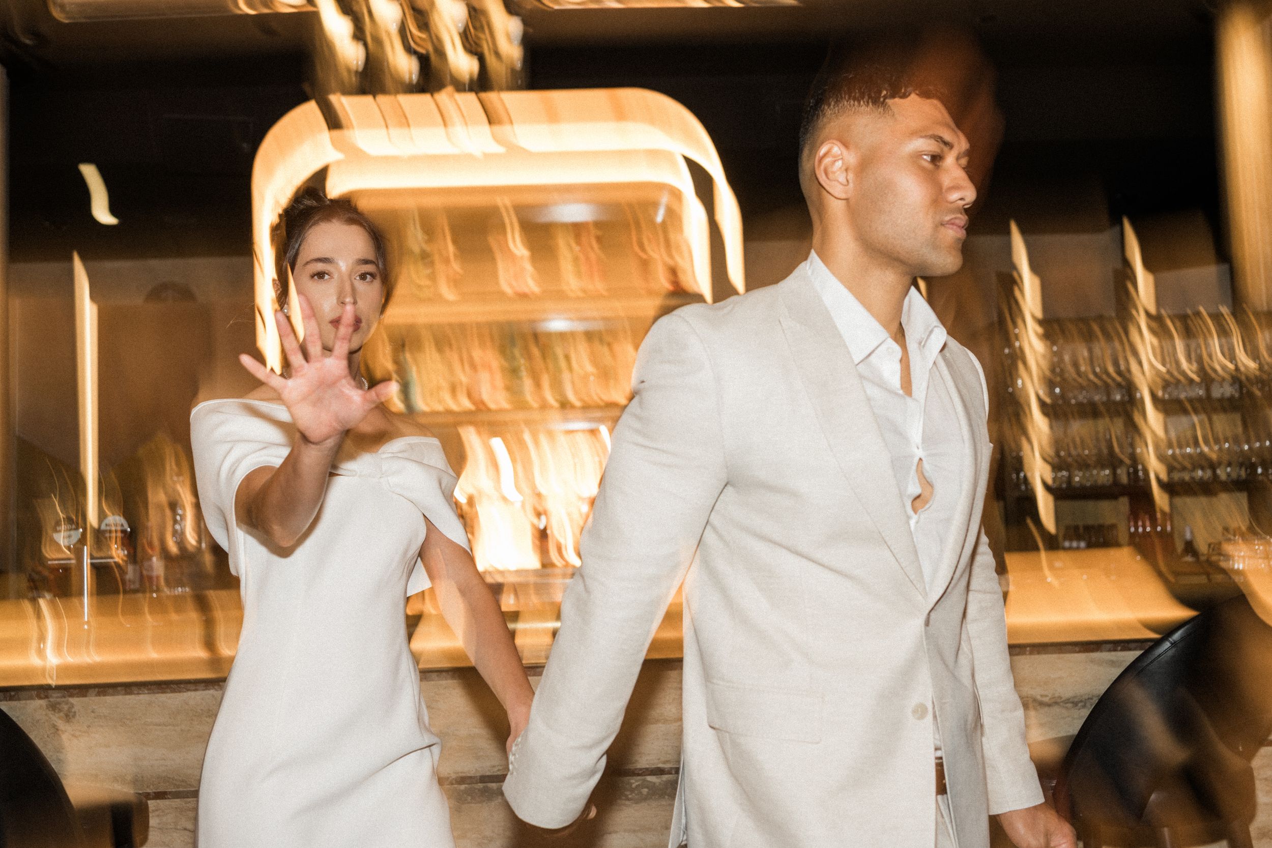A woman in white waves her hand while a man in a white suit stands beside her in an elegant interior setting.