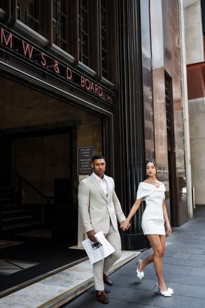 Elegant couple in white mini dress and beige suit holding hands outside a classic city building.