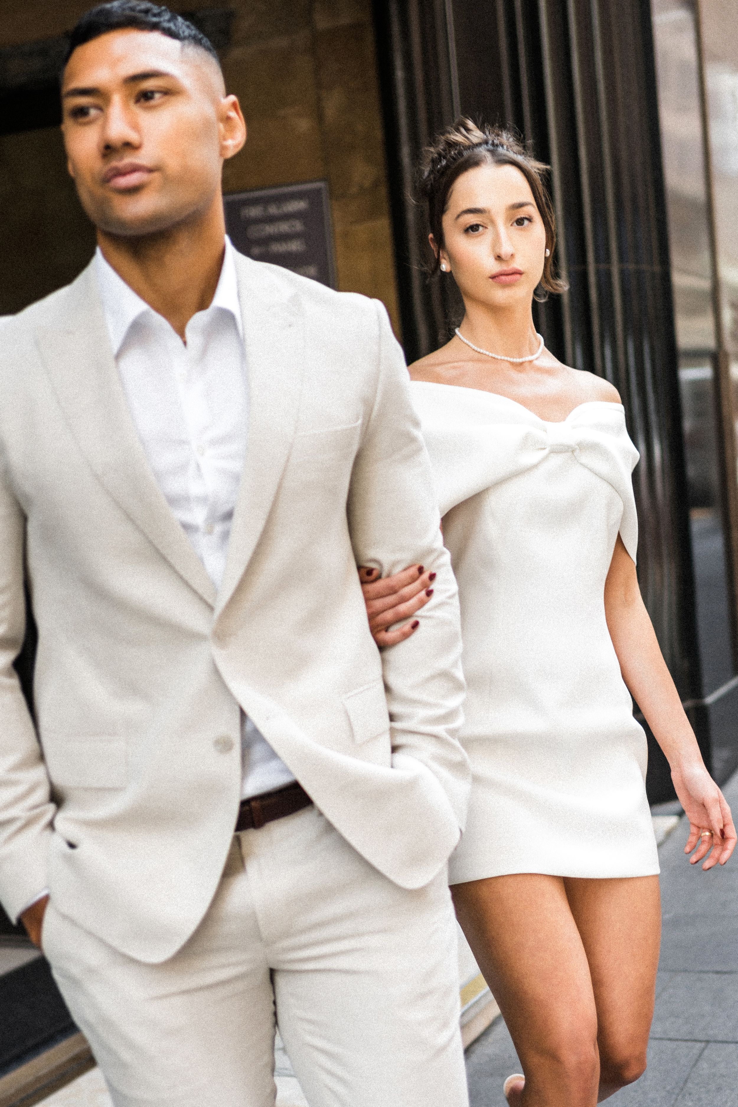 Couple in matching ivory outfits, man in suit and woman in off-shoulder mini dress, walking elegantly outdoors.