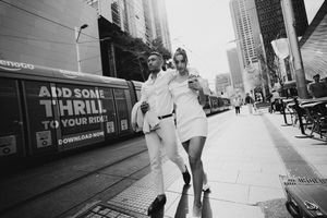 Stylish couple in white outfits walking on a city street in black and white photo with a tram in background.
